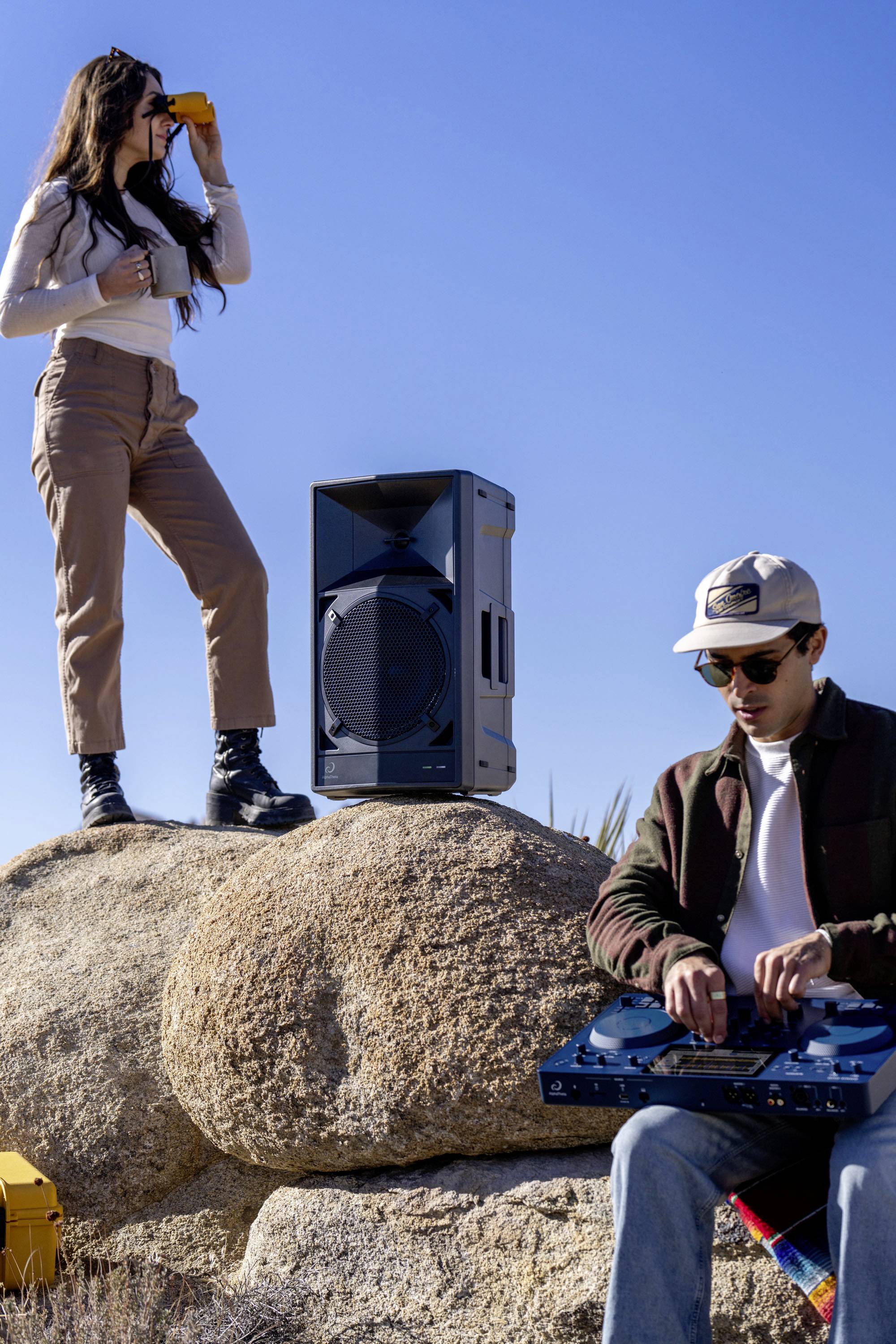 Une femme avec des jumelles se tient sur un rocher à côté d'un grand haut-parleur. Un homme manipule une table de mixage DJ sur un rocher adjacent.