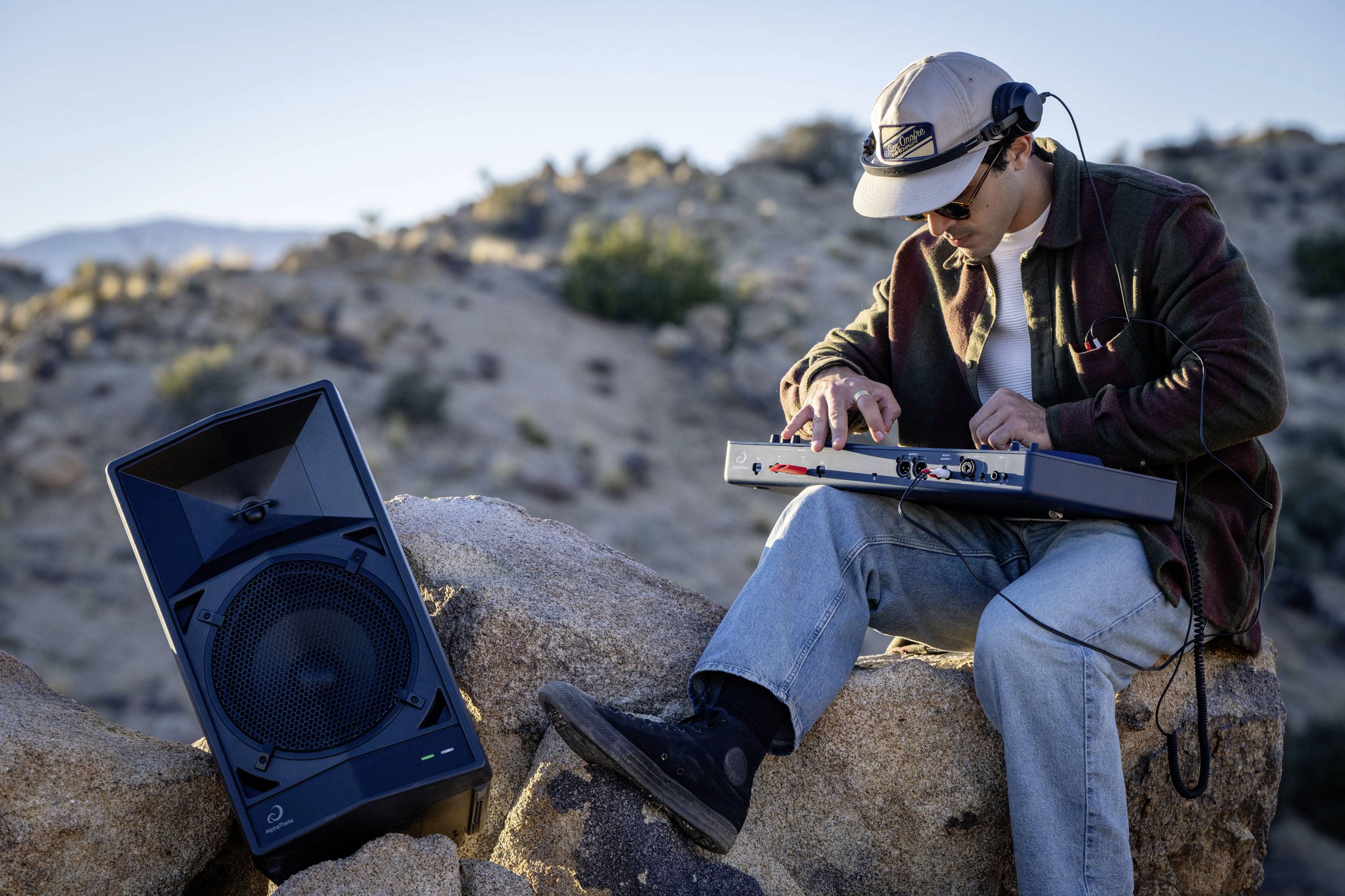 Une personne est assise sur un rocher dans le désert et joue un appareil musical portable, avec un haut-parleur à ses côtés.