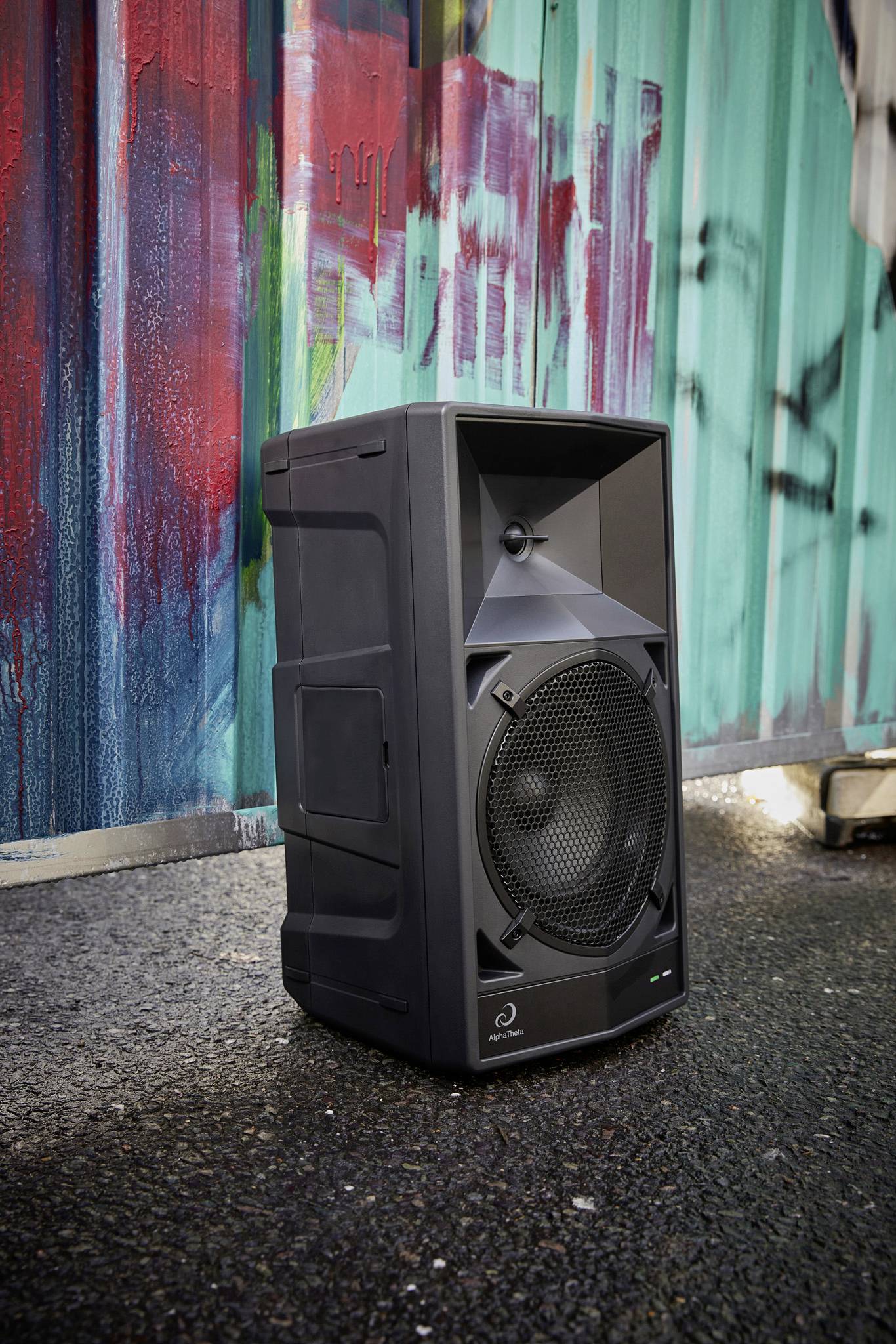 Un grand haut-parleur noir est posé sur le sol devant un mur coloré recouvert de graffitis. L'emplacement semble urbain.