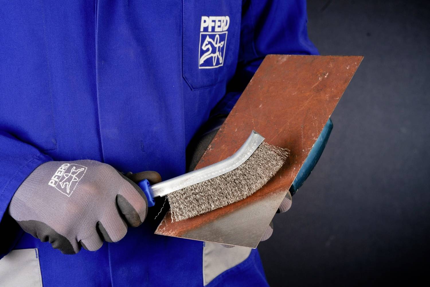 Une personne en vêtements de travail bleus nettoie une tôle métallique avec une brosse métallique.