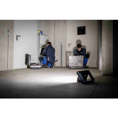 Deux personnes en tenue de travail travaillent dans une salle technique. Une personne vérifie un équipement électrique, tandis que l'autre trie des caisses.