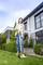 Une femme tond la pelouse avec une tondeuse électrique devant une maison moderne. Il fait soleil et le jardin est bien entretenu.