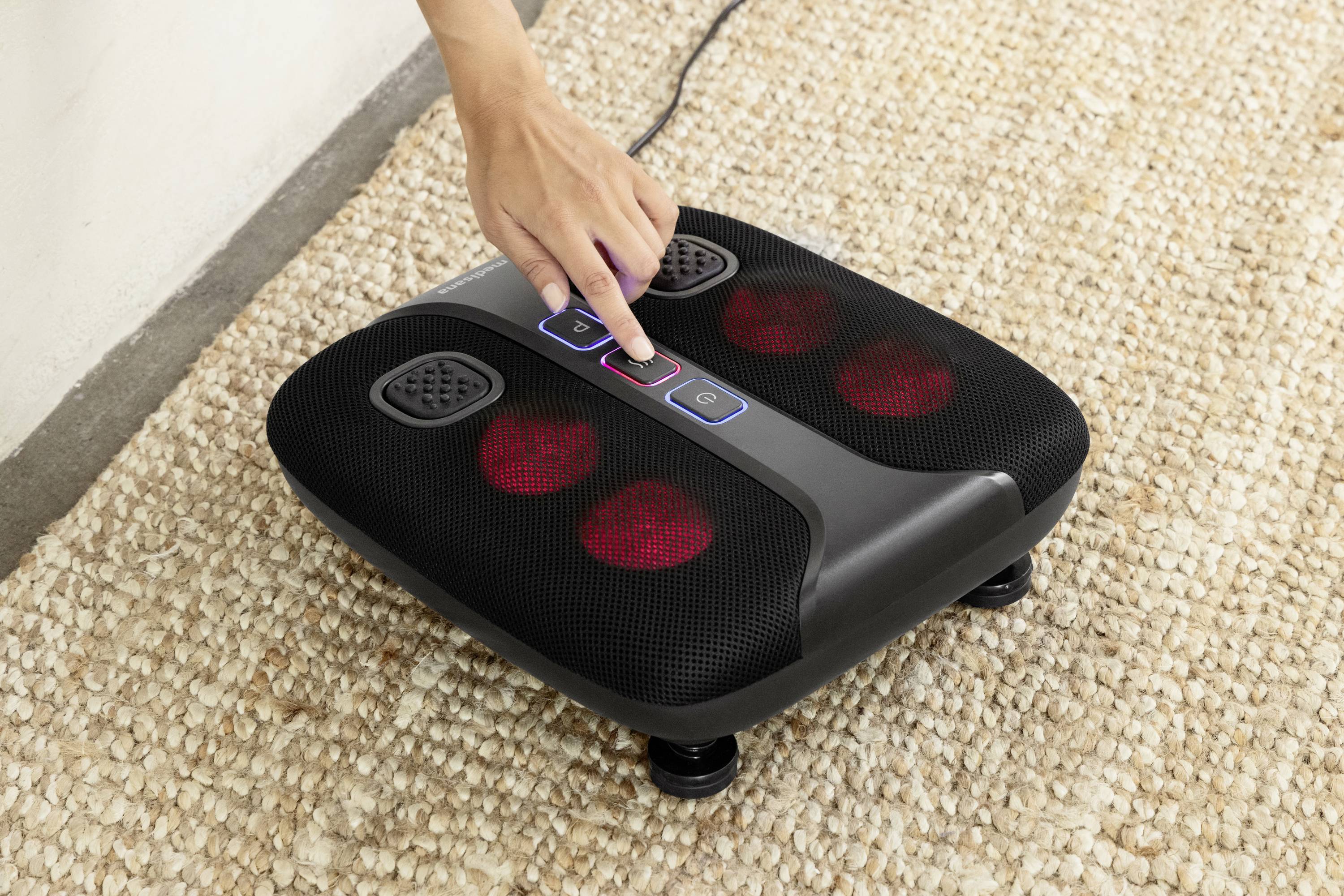 Un appareil de massage pour les pieds est utilisé sur un tapis. Une main appuie sur un bouton de l'appareil, qui fonctionne avec des lumières brillantes.