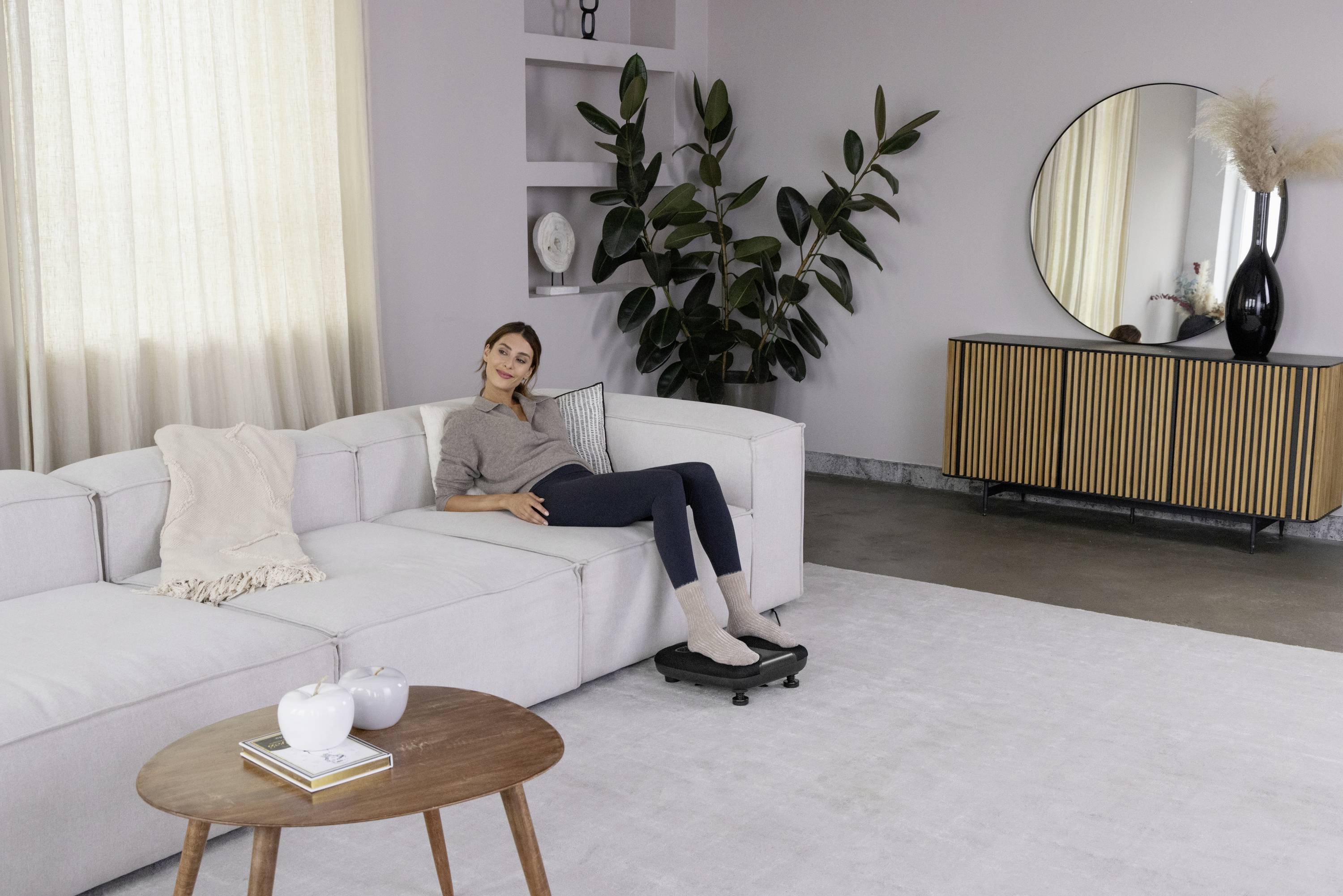 Une femme est allongée de manière détendue sur un canapé blanc dans un salon moderne, avec des plantes, un miroir rond et une armoire en bois en arrière-plan.