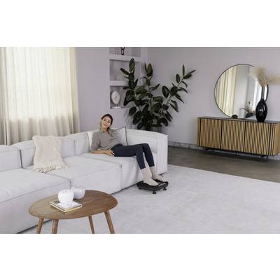 Une femme est allongée de manière détendue sur un canapé blanc dans un salon moderne, avec des plantes, un miroir rond et une armoire en bois en arrière-plan.