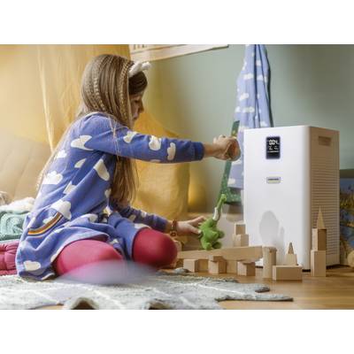 Une fille en robe bleue joue avec des blocs de bois à côté d'un purificateur d'air dans la chambre d'enfant. Le purificateur d'air affiche '02%' sur son écran.