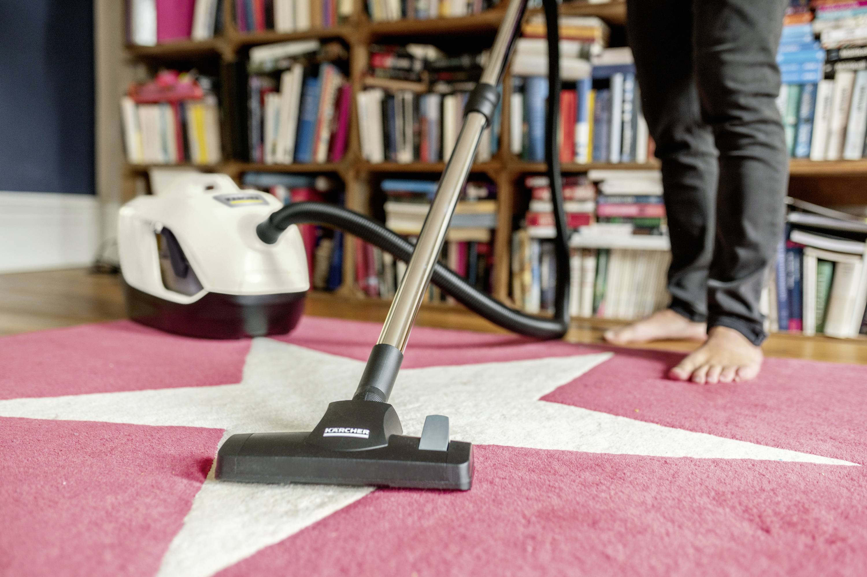 Une personne aspire un tapis rouge à motif d'étoiles blanches devant une bibliothèque. L'accent est mis sur l'aspirateur.
