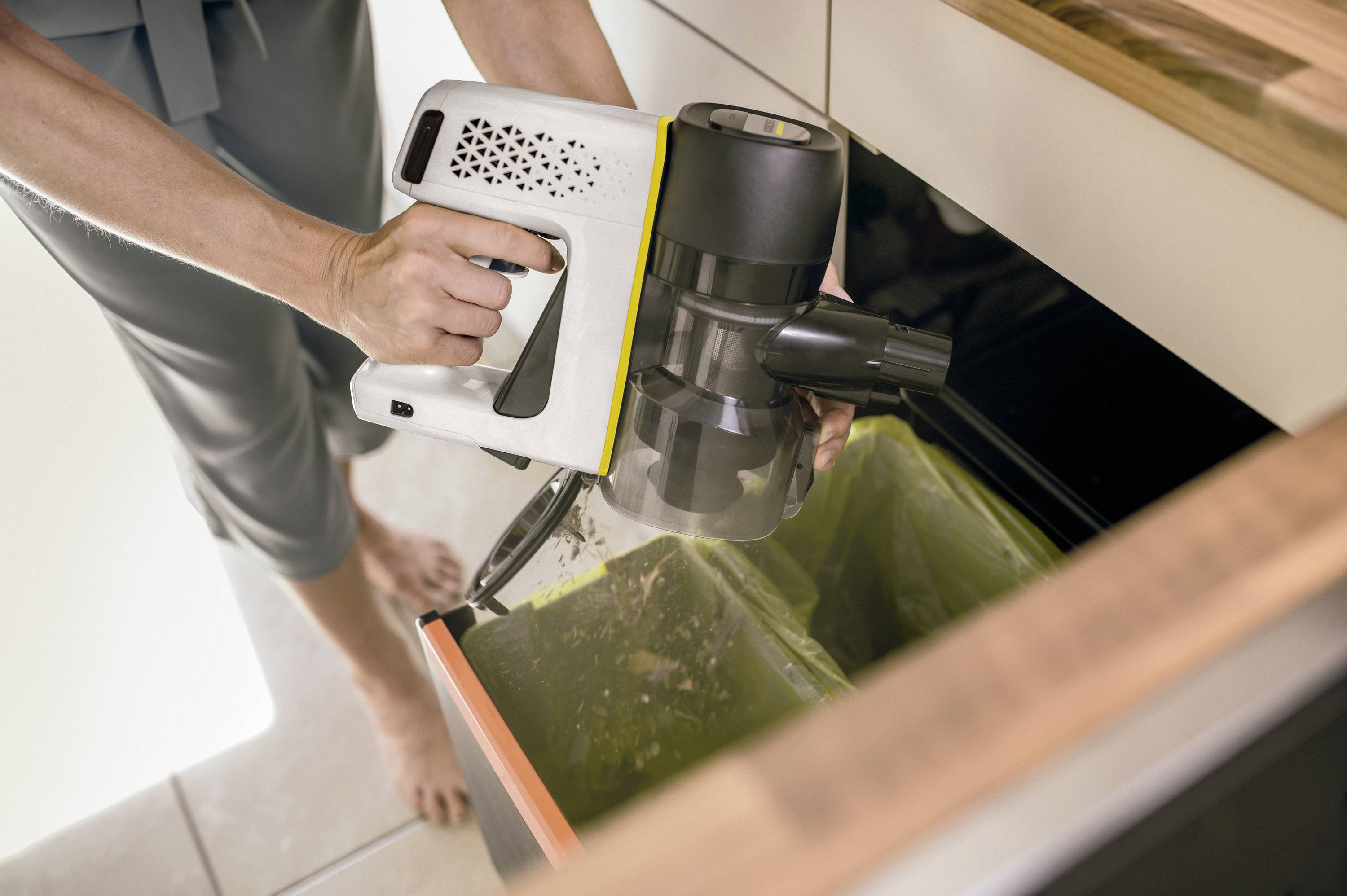 Une personne vide le contenu d'un aspirateur sans fil dans une poubelle de cuisine. Les mains et l'aspirateur sont visibles.
