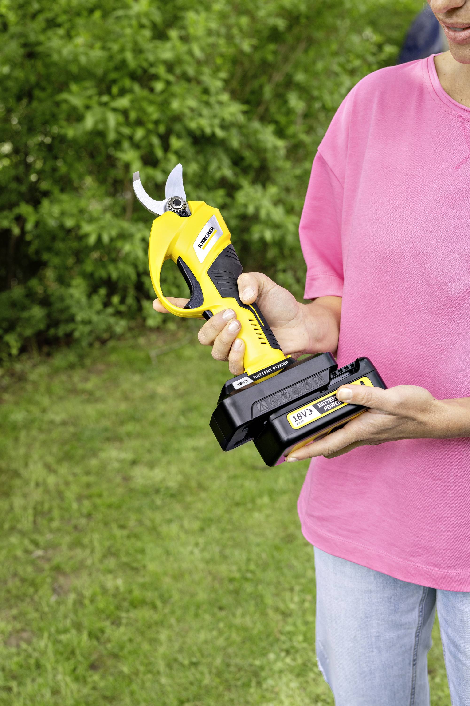 Une personne portant un t-shirt rose tient un sécateur électrique jaune avec sa batterie installée, debout sur une pelouse.