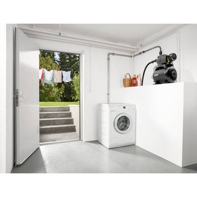 Salle de lavage avec une machine à laver blanche, à côté de laquelle se trouve une pompe sur un piédestal. À l'arrière-plan, une porte mène à un jardin avec une corde à linge.