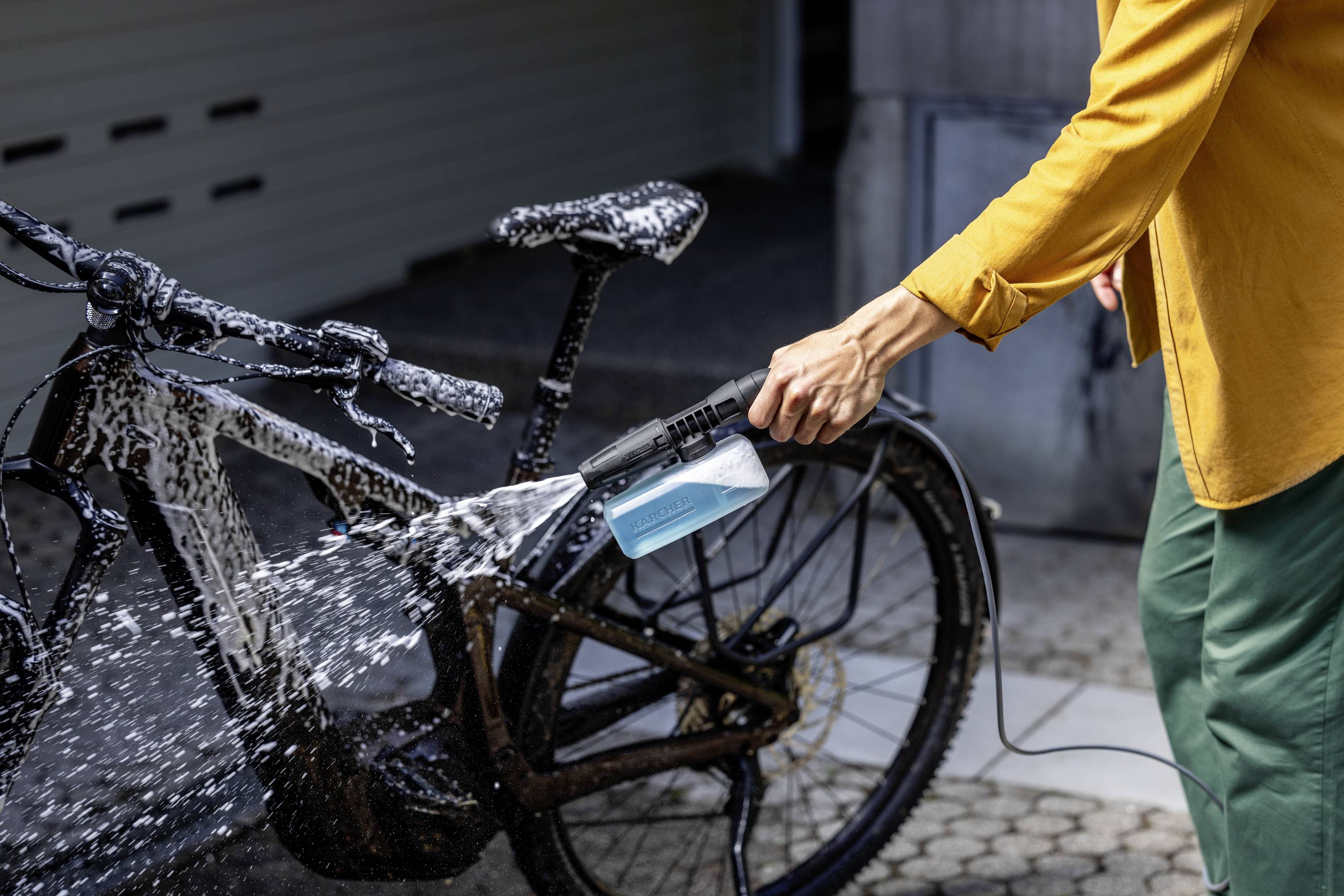 Une personne lave un vélo avec un nettoyeur haute pression, le vélo est couvert d'eau savonneuse.