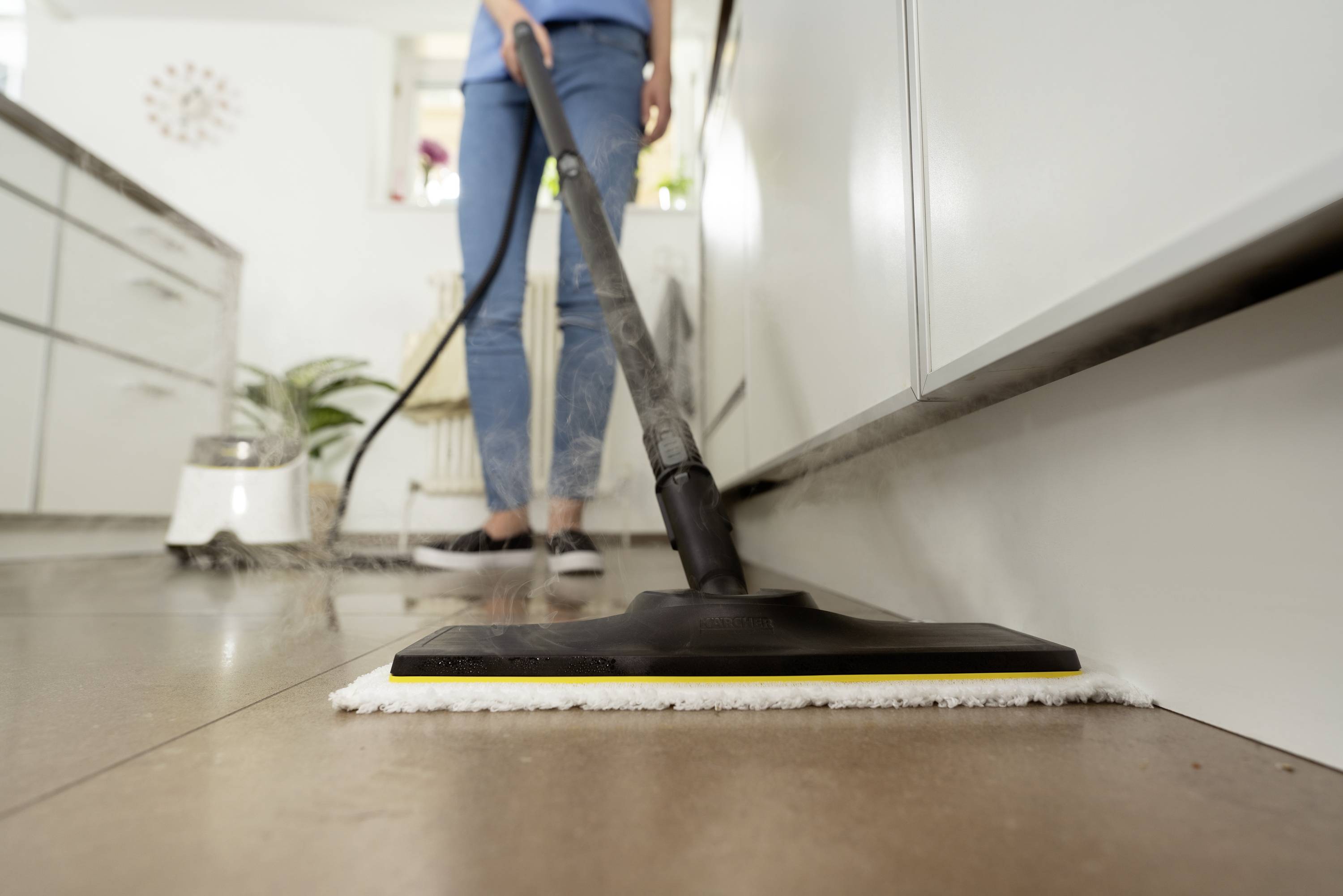 Une personne nettoie le sol de la cuisine avec un nettoyeur vapeur. Des placards de cuisine et une fenêtre sont visibles en arrière-plan.