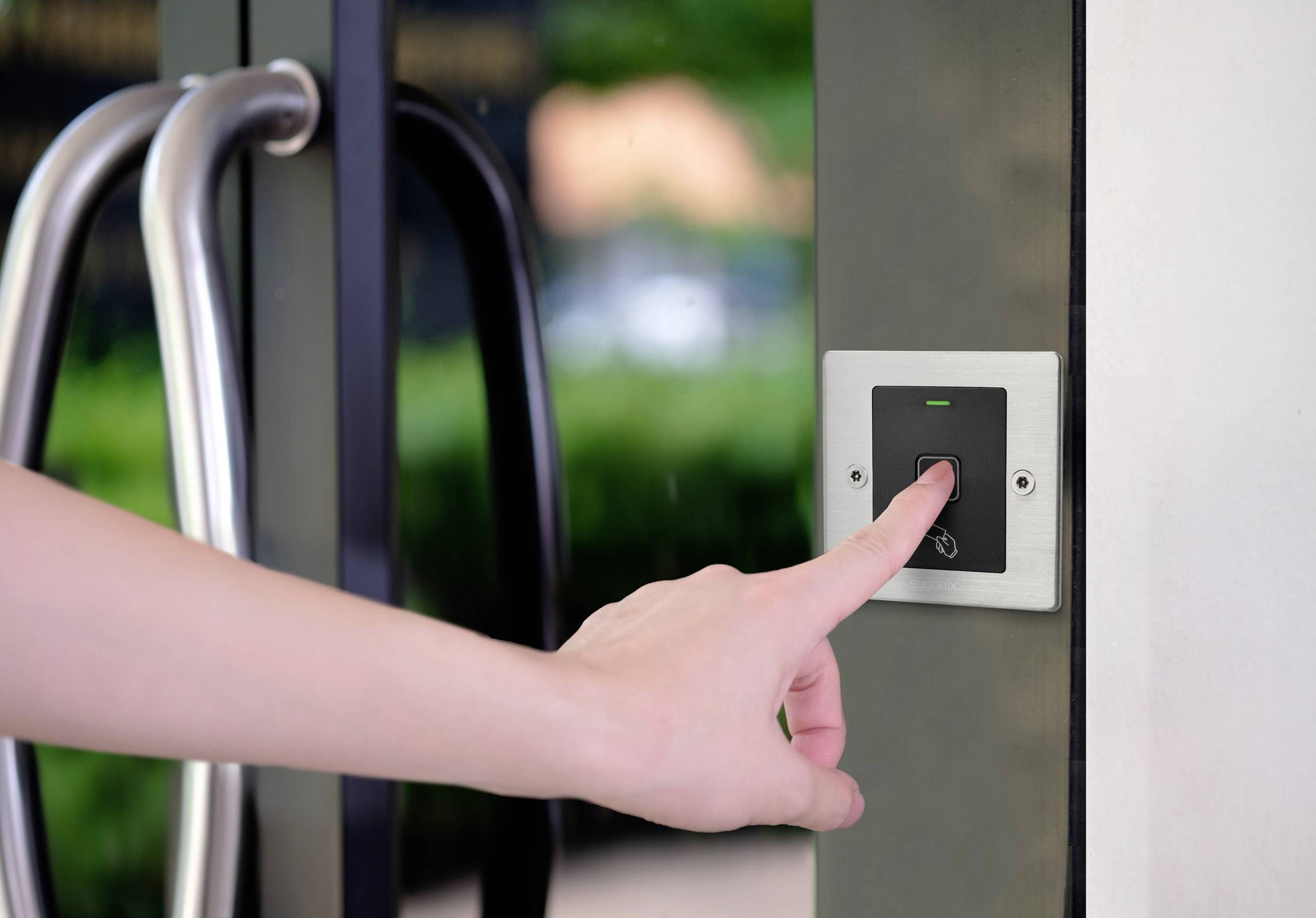 Une main appuie sur un bouton d'accès à côté d'une porte en verre, le voyant vert s'allume. En arrière-plan, des plantes sont visibles de manière floue.