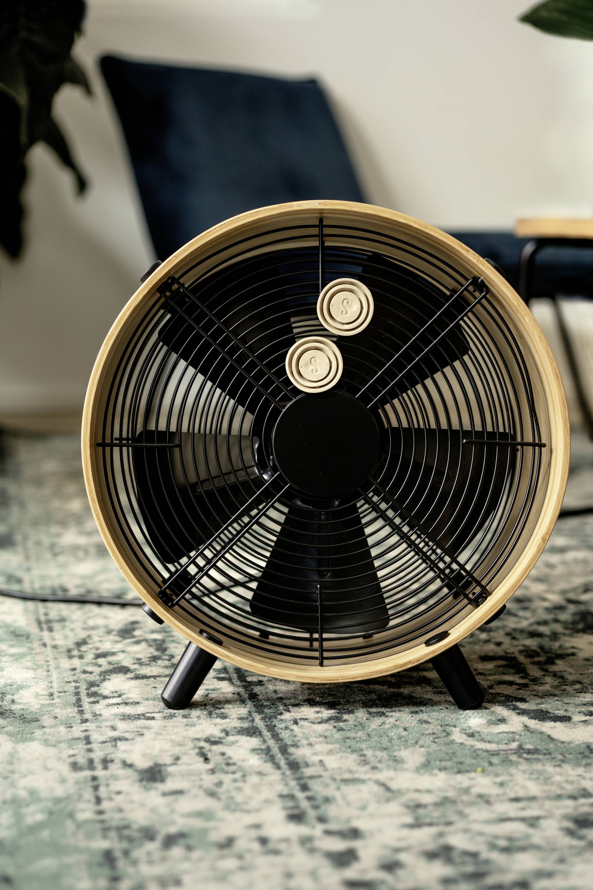 Un ventilateur de table en bois est posé sur un tapis à motifs devant une chaise bleue. Le ventilateur a un design moderne.