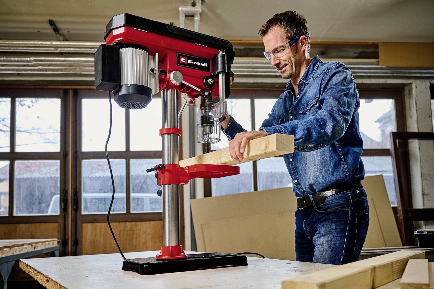 Un homme travaille une pièce de bois avec une perceuse à colonne rouge dans un atelier. Il porte des lunettes de protection et des vêtements en jean.