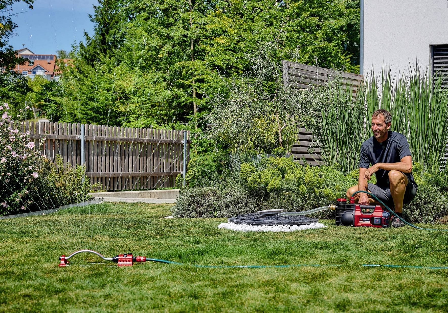 Un homme est agenouillé dans le jardin près d'une pompe à eau, tandis qu'un arroseur irrigue l'herbe. Des arbres et une clôture sont visibles en arrière-plan.