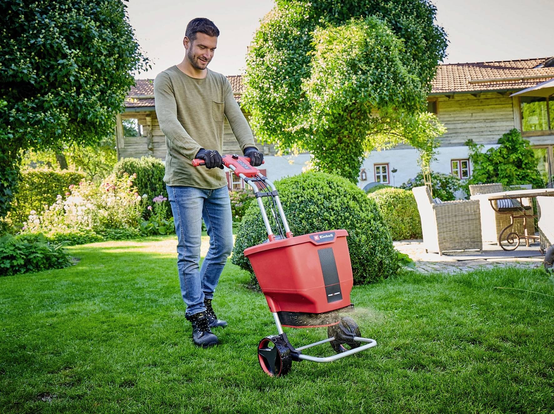 Un homme pousse un chariot d'épandage rouge sur la pelouse dans le jardin. En arrière-plan, on aperçoit une maison et des buissons.