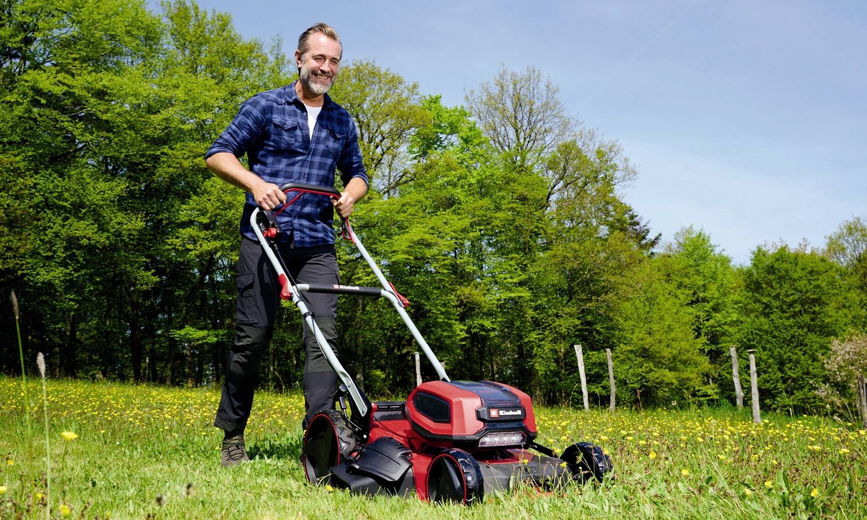 Un homme tond la pelouse avec une tondeuse électrique sur un terrain herbeux par une journée ensoleillée, entouré d'arbres.