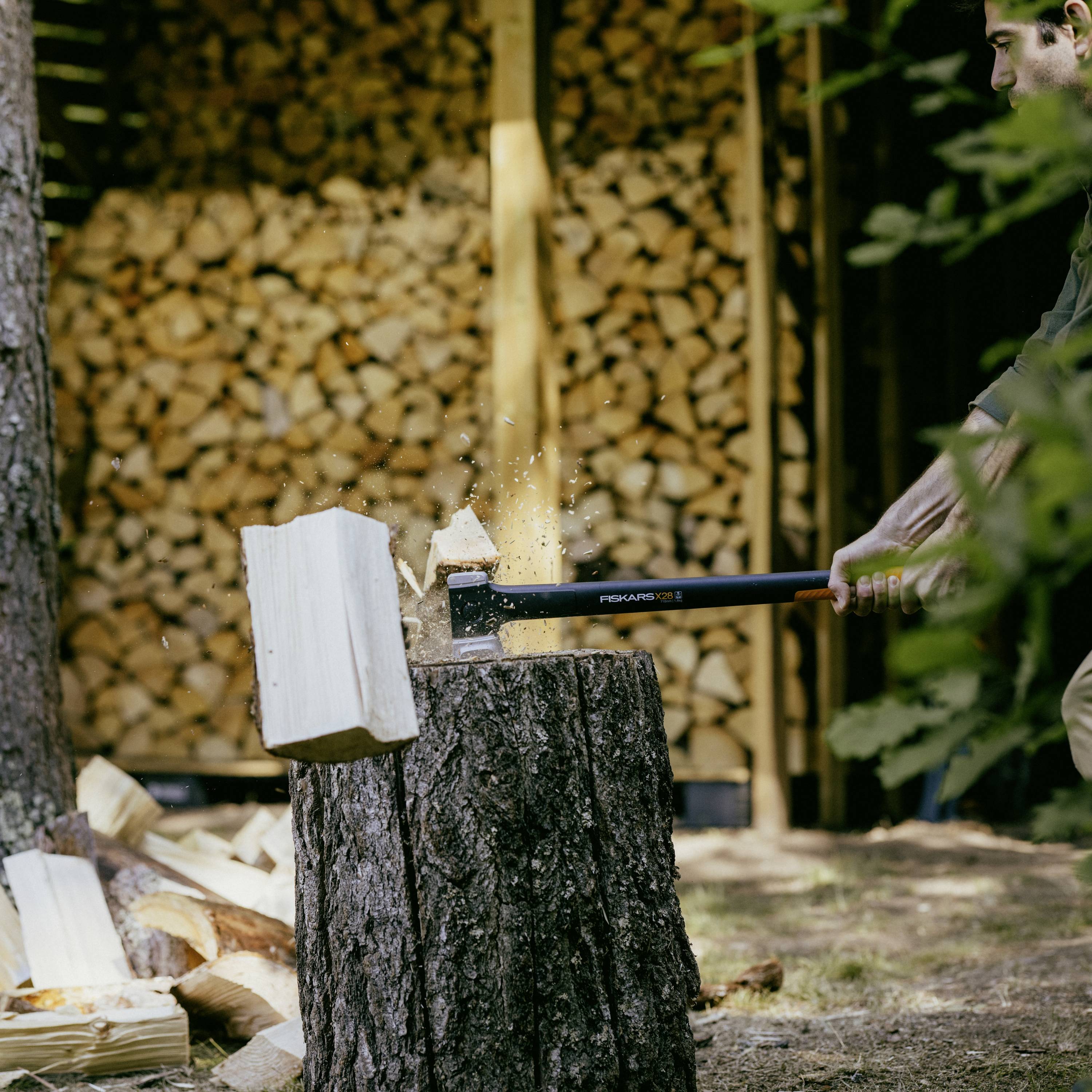 Une personne fend un billot de bois avec une hache sur une souche d'arbre. À l'arrière-plan, on peut voir des tas de bois empilés.