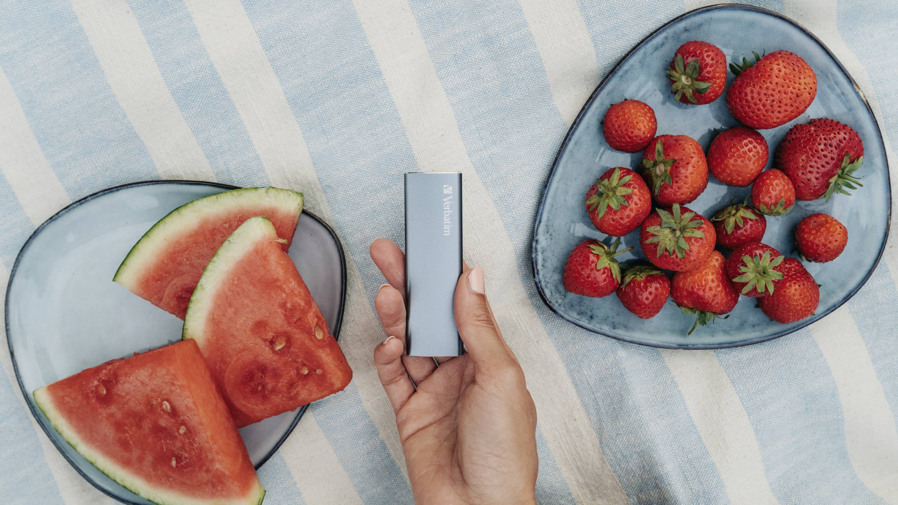 Une main tient un objet rectangulaire bleu. À côté, des tranches de pastèque et de fraises sont disposées sur de la vaisselle bleue.