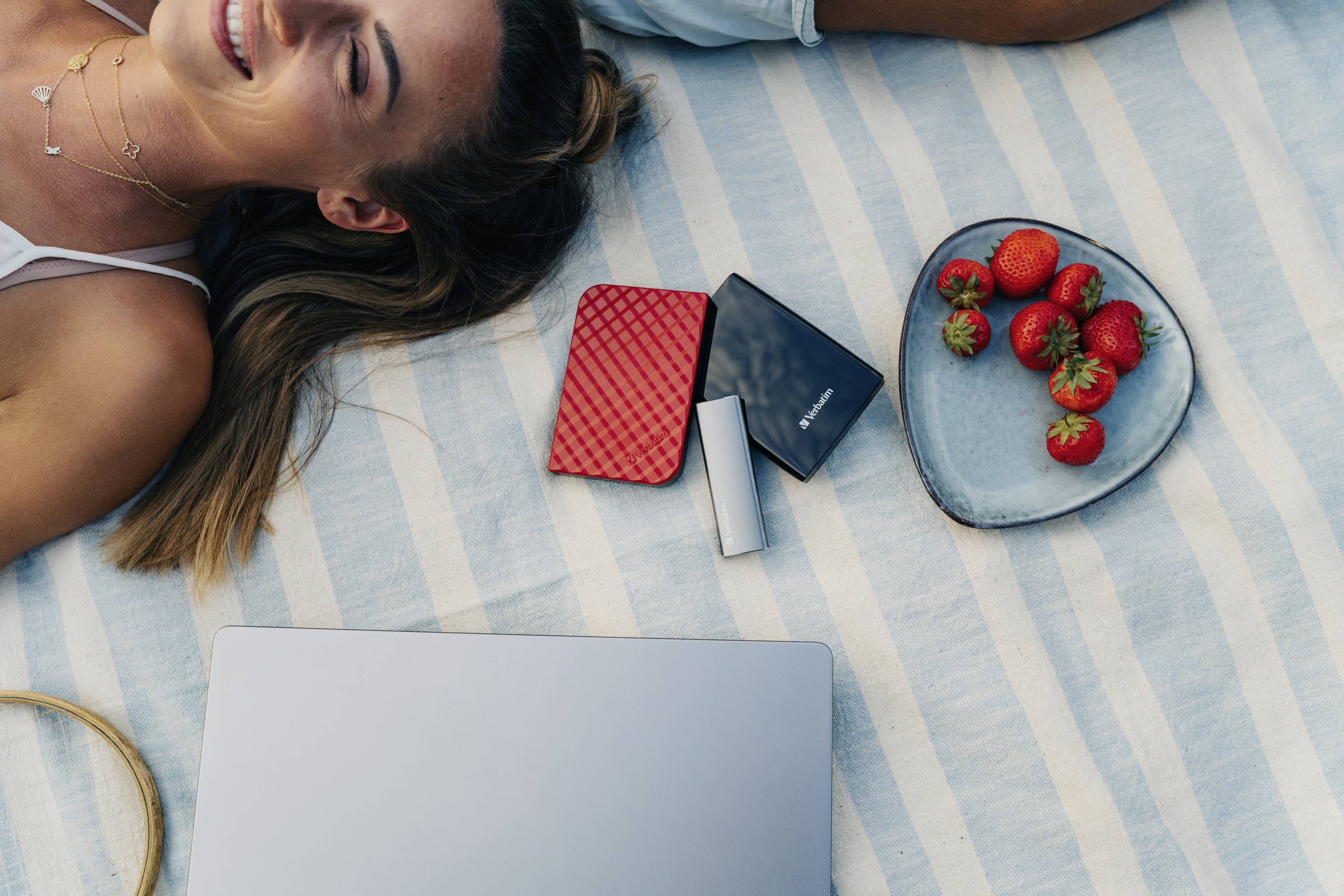 Une femme est allongée, souriante, sur une couverture de pique-nique avec des disques durs externes et un plat de fraises à côté d'elle. Un ordinateur portable est posé à proximité.