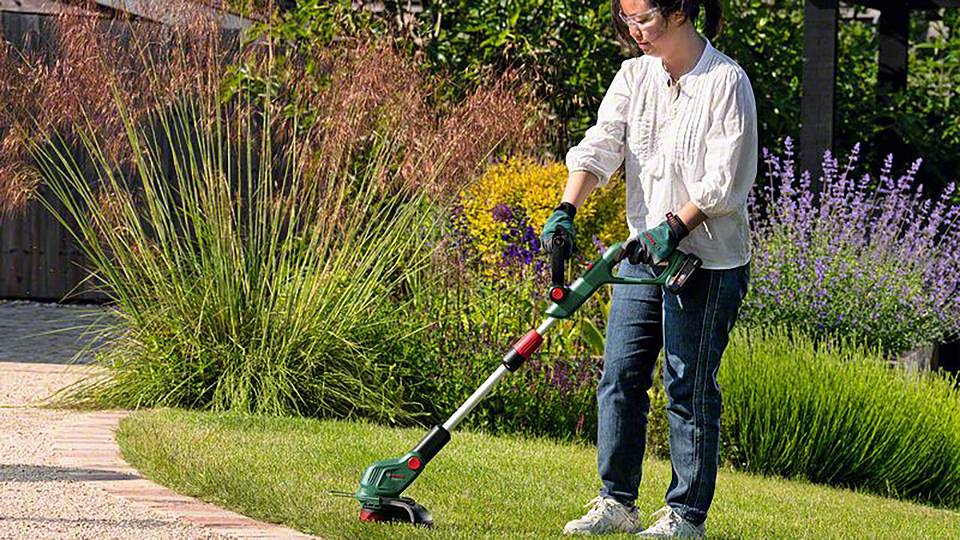 Une personne tond la pelouse avec un coupe-bordure électrique dans le jardin. Entourée de fleurs colorées et de buissons verts. Le temps est ensoleillé.