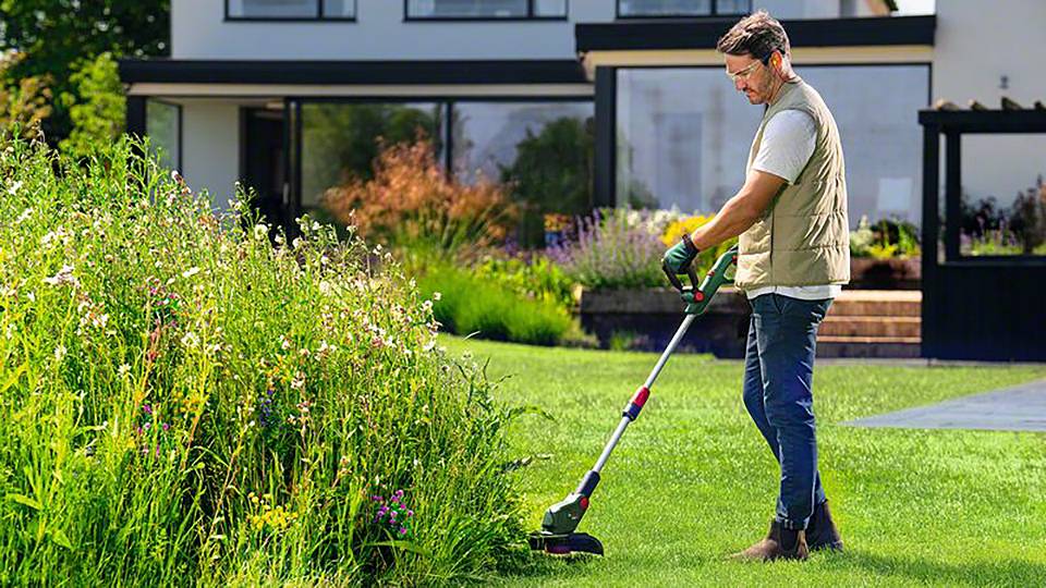 Un homme utilise un coupe-bordure pour tailler l'herbe au bord d'un jardin bien entretenu. À l'arrière-plan se trouve une maison moderne.