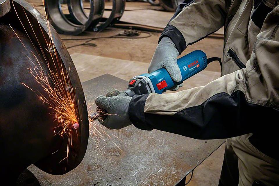 Un ouvrier utilise une meuleuse d'angle pour travailler le métal. Des étincelles jaillissent au point de contact. Des vêtements de protection et des gants sont portés.