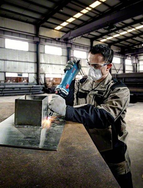 Un artisan en vêtements de protection travaille une pièce métallique dans un atelier. Il utilise un outil électrique pour poncer.