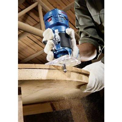 Une personne fraise un bord de bois avec un appareil de fraisage bleu. Des copeaux de bois volent pendant que le bord est usiné.