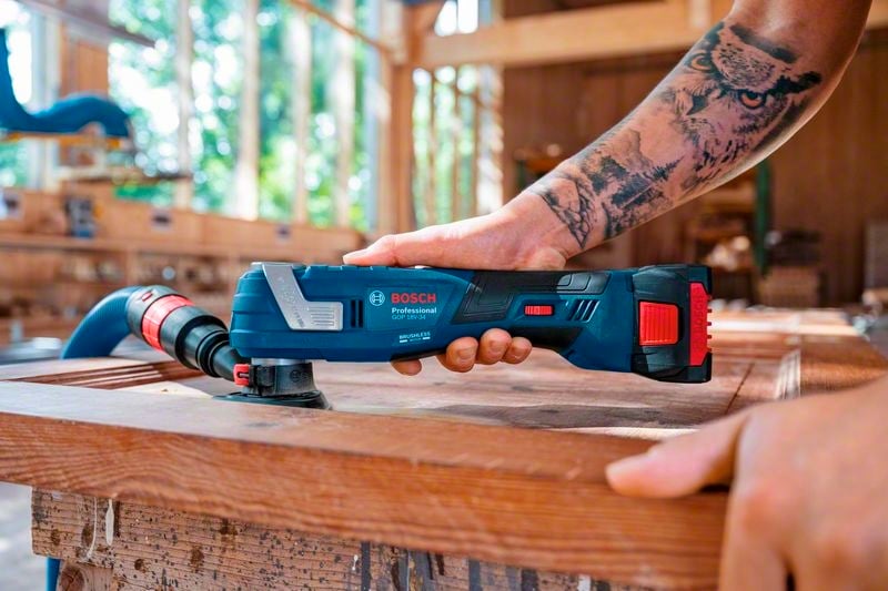 Une personne utilise un outil multifonction électrique pour poncer du bois dans un atelier. Des tatouages sont visibles.