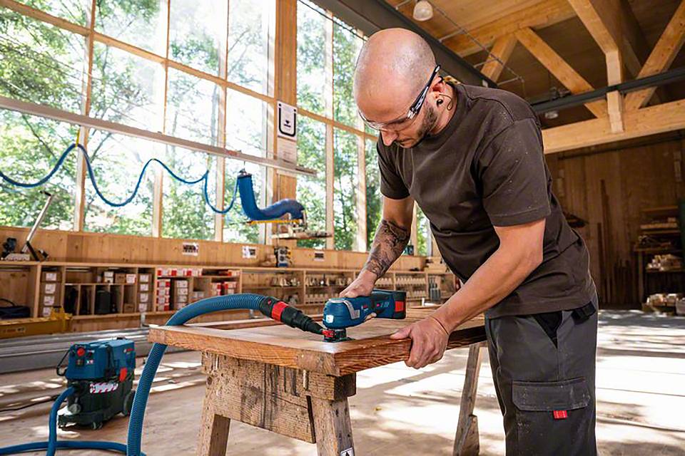 Un homme ponce un morceau de bois dans un atelier baigné de lumière naturelle. Il porte des lunettes de protection et des protections auditives et utilise un outil électrique.