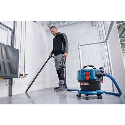 Un homme se tient dans une pièce de sous-sol et nettoie le sol avec un aspirateur eau et poussière. La pièce a des murs blancs et différentes canalisations.