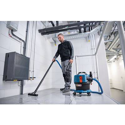 Un homme nettoie le sol d'une salle technique à l'aide d'un aspirateur industriel. Des tuyaux et un boîtier de distribution sont visibles sur le Mur.