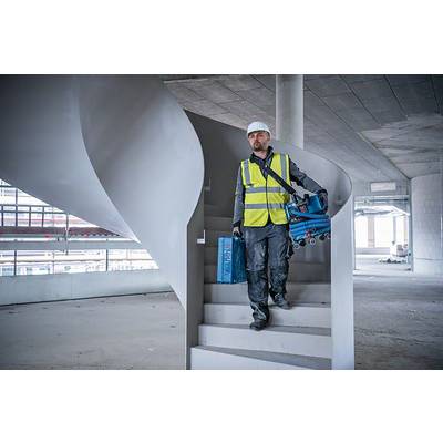 Un ouvrier portant un casque de chantier et un gilet de sécurité descend un escalier dans un bâtiment en construction, des outils à la main.