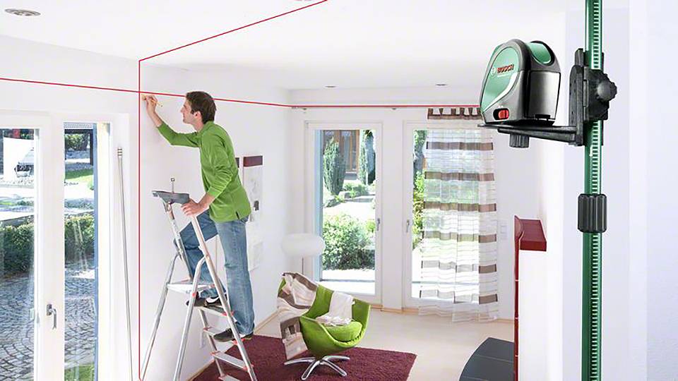 Un homme mesure avec un appareil de nivellement laser projetant des lignes rouges au plafond, alors qu'il travaille sur une échelle dans un salon lumineux.