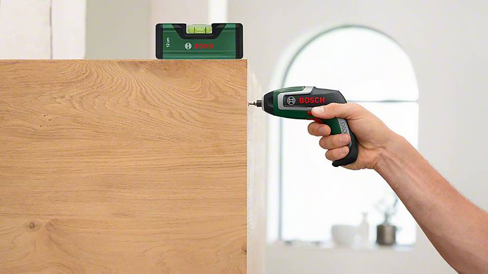 Une main tient une visseuse sans fil Bosch et visse dans du bois. Au-dessus, un niveau à bulle Bosch vert est posé verticalement.