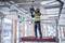 Un travailleur portant un gilet de sécurité et un casque installe des équipements électriques au plafond d'un chantier, entouré d'échafaudages et de tuyaux.