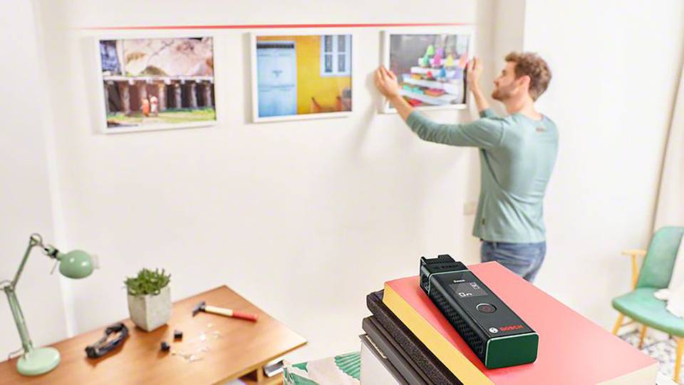 Un homme accroche un tableau au mur. Au premier plan, un télémètre laser est posé sur une pile de livres. Dans la pièce se trouvent une table et une lampe.
