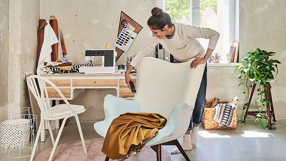 Une femme installe une chaise dans un bureau lumineux et créativement aménagé, avec une machine à coudre, des tissus et des plantes.