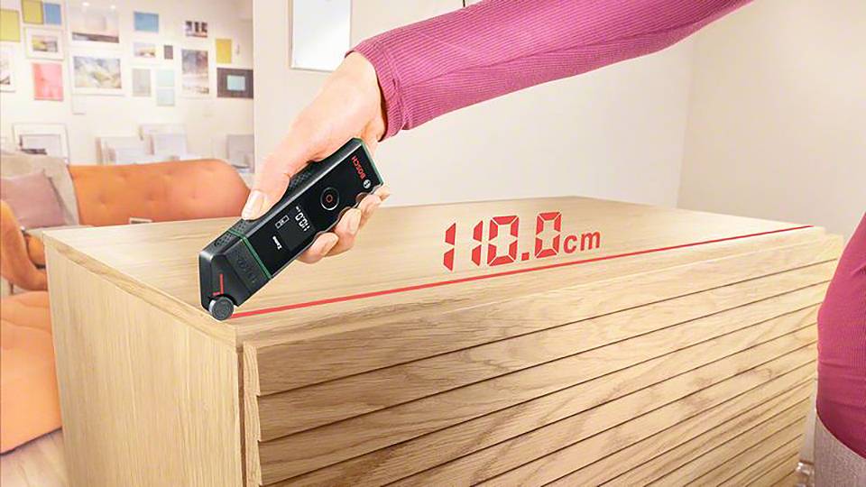 Une personne mesure la largeur d'une caisse en bois à l'aide d'un télémètre laser numérique. L'écran affiche '180,0 cm'.