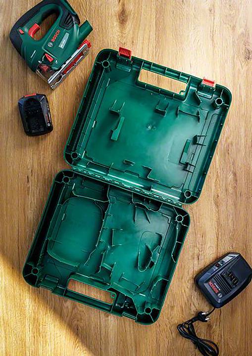 'Boîte à outils verte ouverte sur un plancher en bois, avec une scie sauteuse, une batterie et un chargeur à côté.'