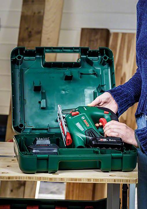 Une personne dépose une scie sauteuse verte dans une boîte à outils ouverte sur une table en bois dans un atelier.
