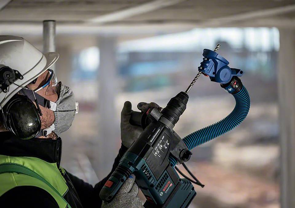 Un ouvrier portant un casque de protection et un masque utilise une perceuse électrique sur un chantier de construction. Il porte des vêtements de sécurité verts.