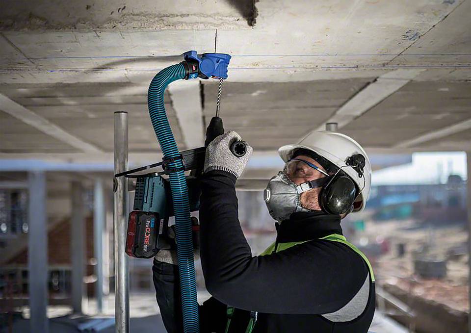 Un ouvrier porte des équipements de protection et fore un trou dans une dalle de béton sur un chantier de construction à l'aide d'une machine.