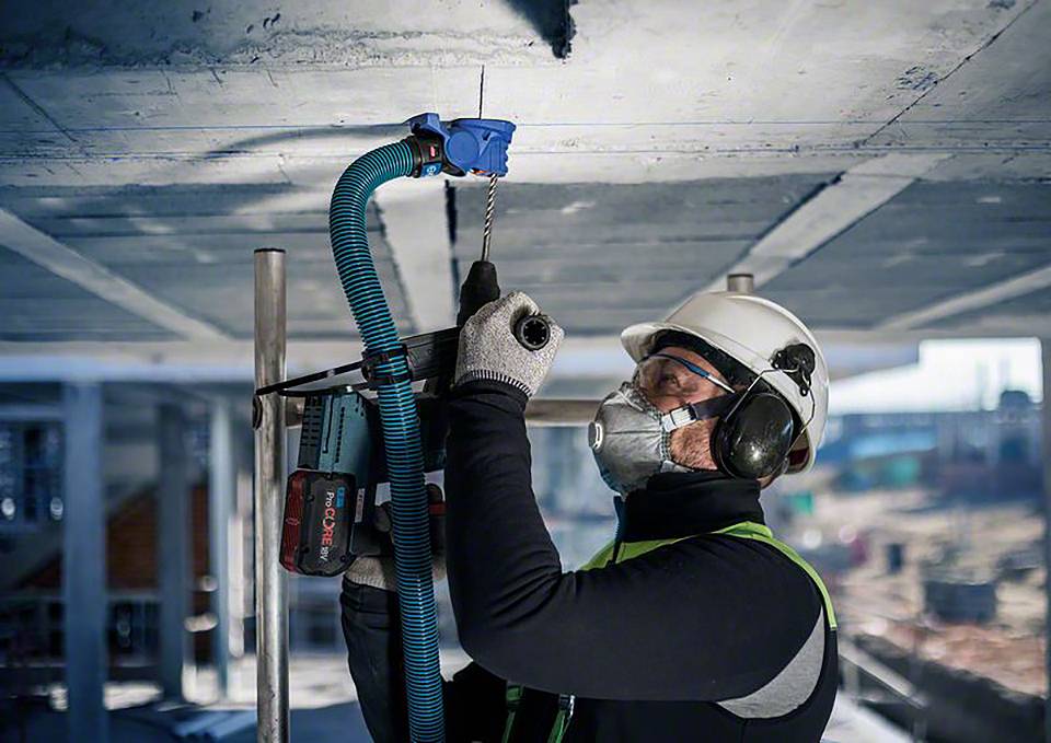 Un ouvrier fore un plafond et porte des équipements de protection, comprenant un casque, des lunettes, un masque et des gants, sur un chantier de construction.