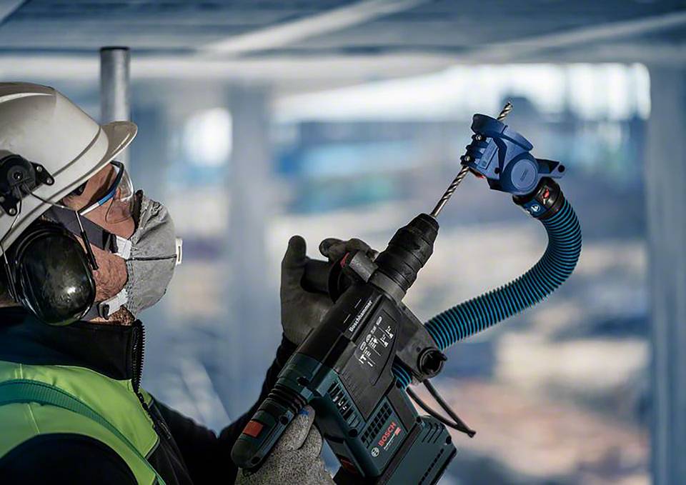 Un ouvrier porte des équipements de protection et fore un poutre en béton à l'aide d'une perceuse. En arrière-plan, on aperçoit un chantier de construction.