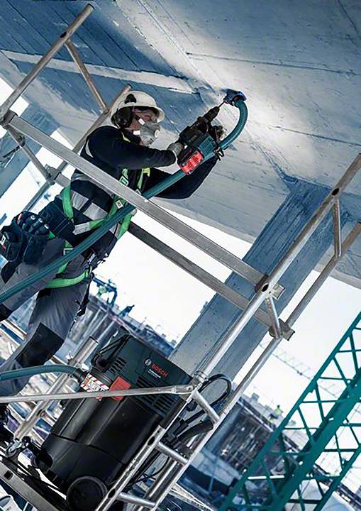Ouvrier sur un échafaudage, portant un équipement de protection, utilisant un outil électrique sur un plafond. En arrière-plan : un chantier de construction avec une grue.