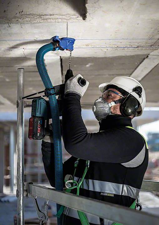 Un ouvrier portant un casque de protection et un masque fore un mur en béton avec une machine sur un chantier de construction.