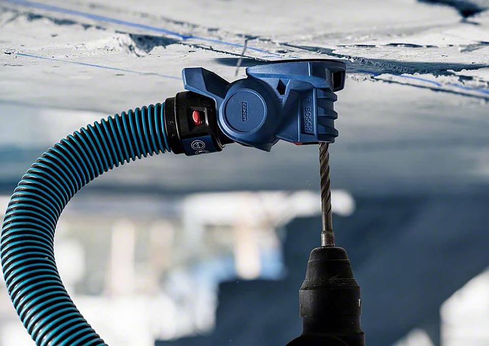 Une tête de forage bleue fixée à une perceuse, perçant une surface. Un tuyau d'aspirateur y est connecté.